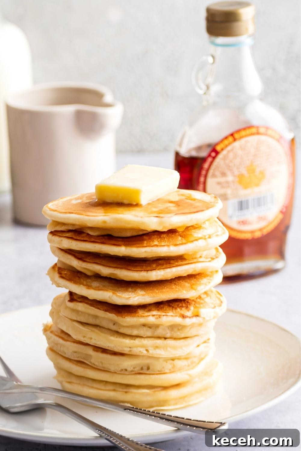 A stack of nine pancakes on a white plate. The top pancake has a square of butter on it. Behind the plate is a bottle of maple syrup.