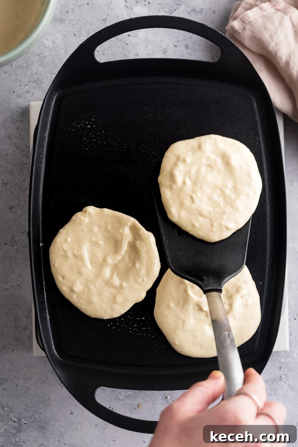 A hand holding a spatula with a pancake on it above a griddle that has two pancakes on it.
