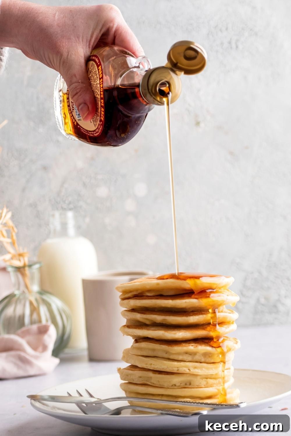 A hand holding a bottle of maple syrup pouring it on a stack of pancakes that are on a white plate with a fork and knife on it.