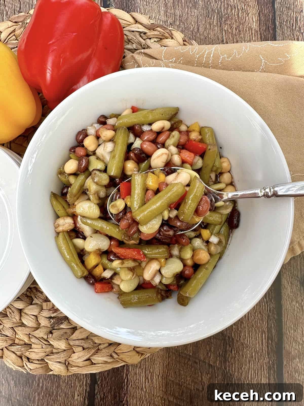 Zesty Mixed Bean Salad with Lemon Herb Dressing 2 A serving bowl of cold green bean salad with a spoon, on a place mat with two peppers off to the side.