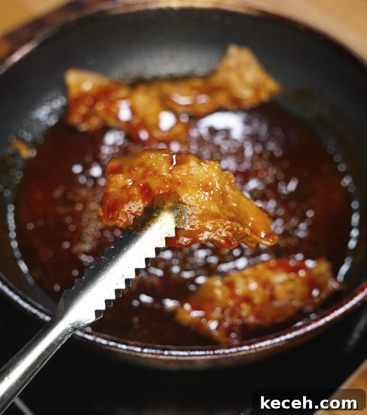 Sticky Spicy Pork Belly Recipe for Crispy Glazed Pork 6 Tongs lifting a slice of fully glazed pork belly from a pan of sticky spicy sauce, showcasing the thick, glossy coating and juicy interior.