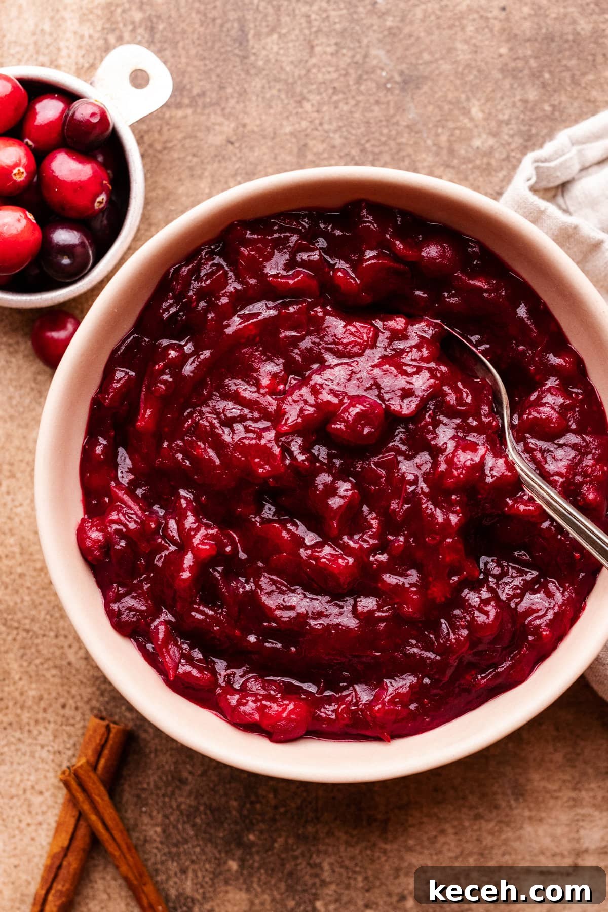 Cranberry Orange Sauce Recipe for Holiday Tables 2 A bowl of cranberry orange sauce with a spoon in it. Around the bowl of sauce is a small bowl of cranberries, cinnamon sticks, and a gray napkin.