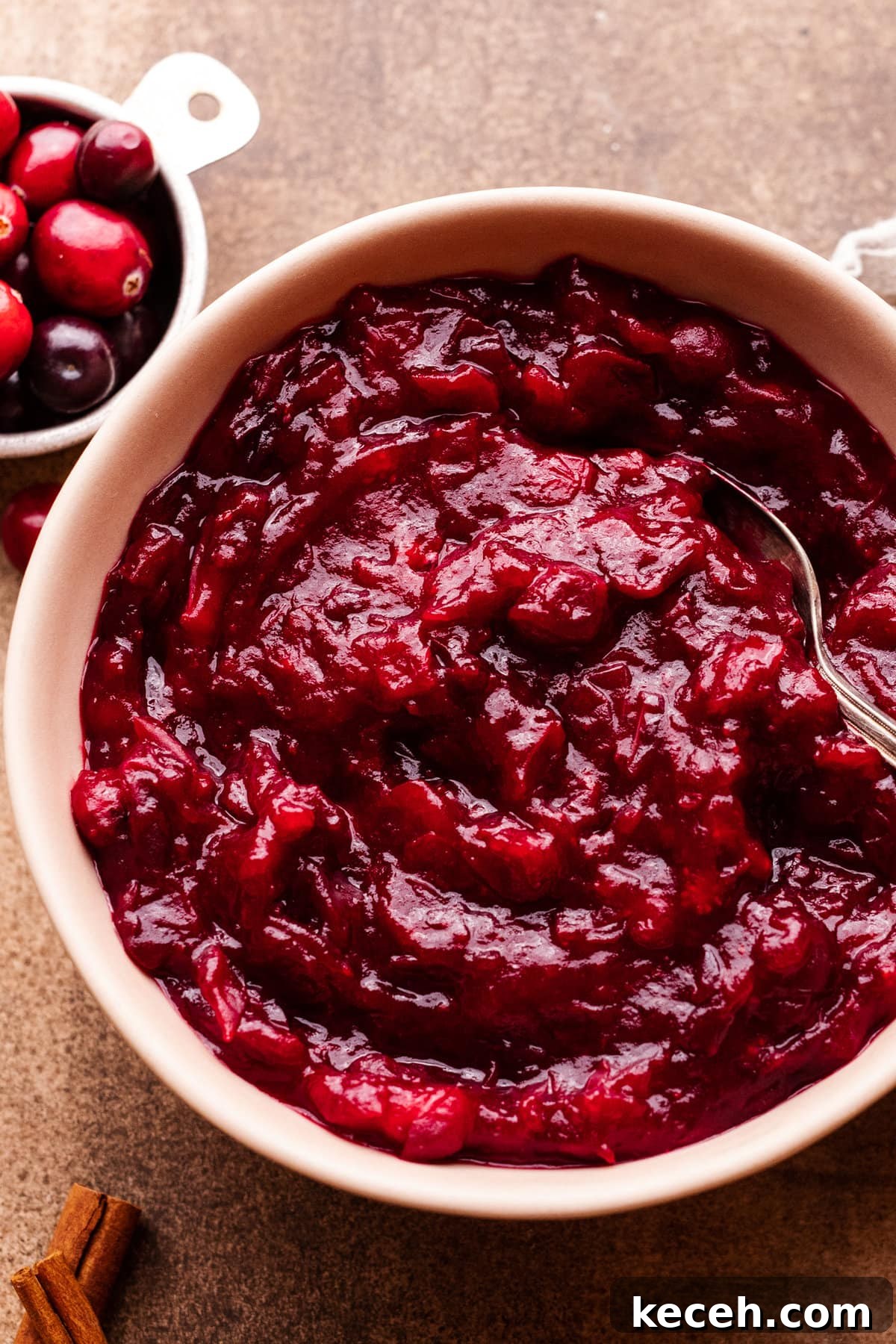 Cranberry Orange Sauce Recipe for Holiday Tables 6 A bowl of cranberry orange sauce with a spoon in it. Around the bowl of sauce is a small bowl of cranberries, cinnamon sticks, and a gray napkin.