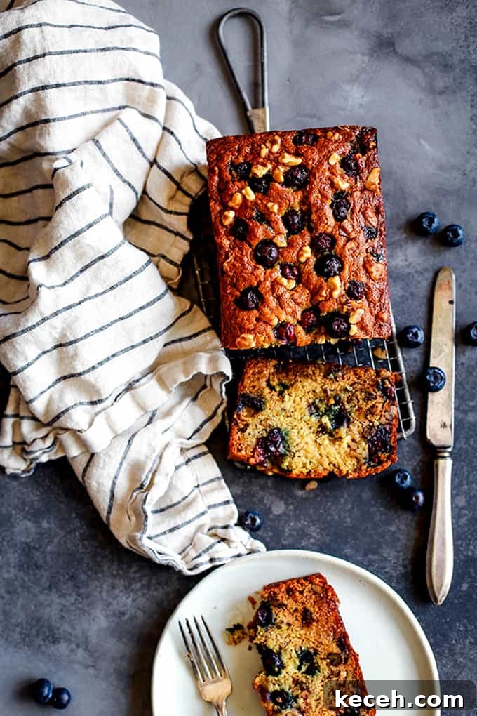 A loaf of Blueberry Walnut Banana Bread flanked by a striped napkin, knife and a plate with a piece of bread on it.