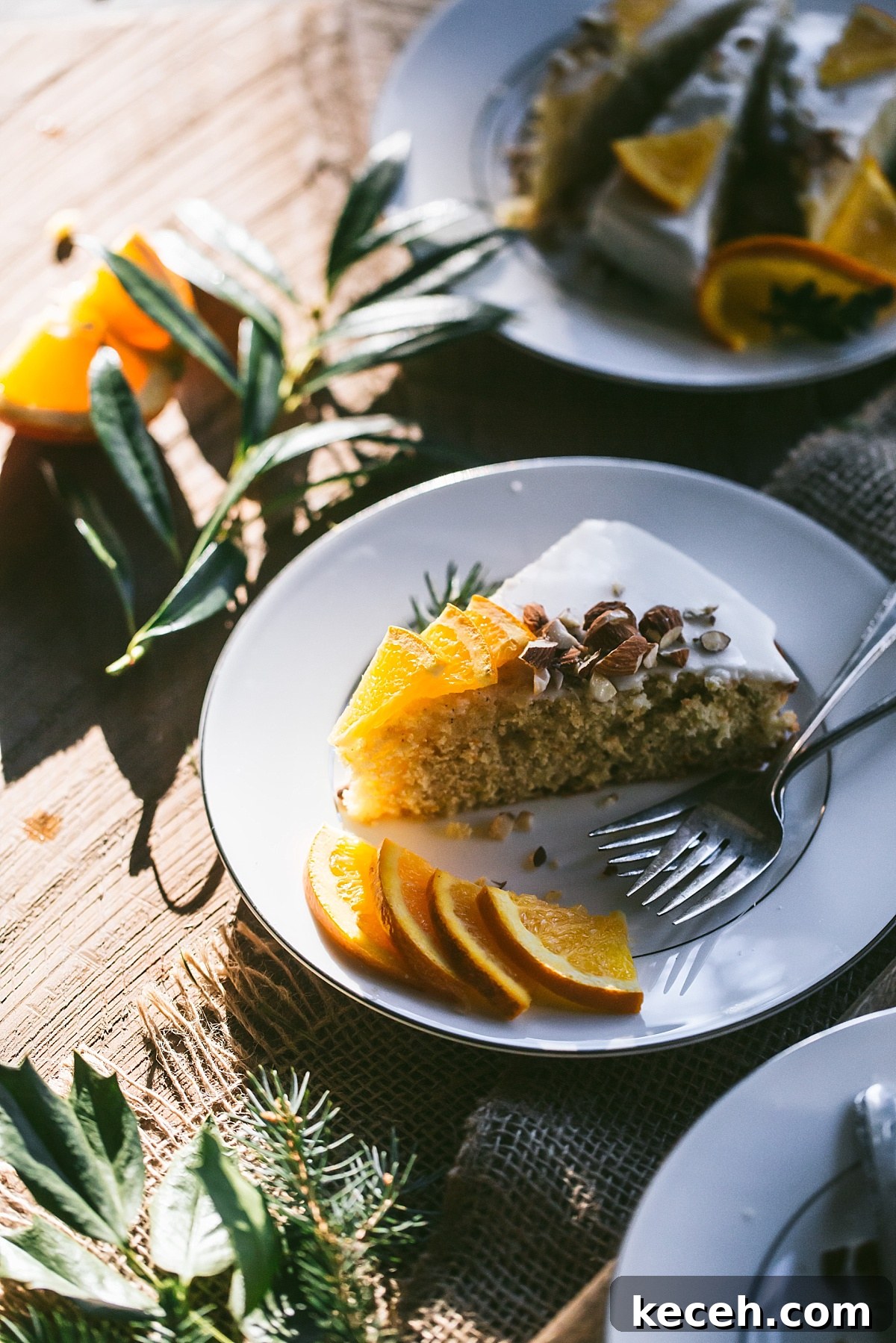 Vasilopita (Greek New Year Cake) — Slow Cooker or Oven Recipe 2 A slice of Vasilopita in sunlight on a white decorative plate topped with sliced oranges and almond pieces.