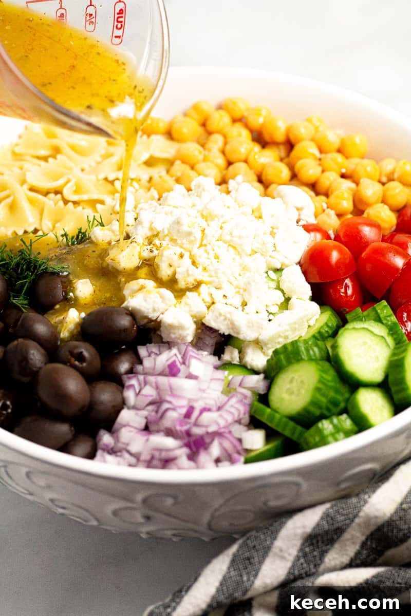 A measuring cup pouring homemade dressing over a white bowl filled with Greek pasta salad