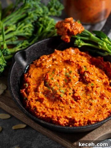 Romesco dip in a small black pan surrounded by broccoli spears, with a bottle of sauce in the background.