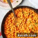Spicy mac and cheese in a large black skillet with a silver spoon inside and pink plate next to it.