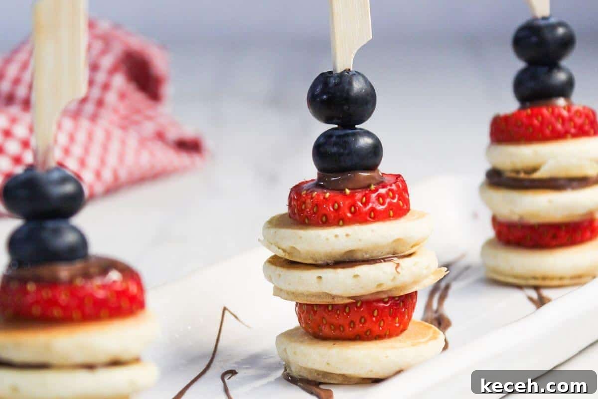 Mini pancake stacks with strawberries, blueberries, and chocolate drizzle on wooden skewers, arranged on a white rectangular plate. A red and white checked cloth is in the background.