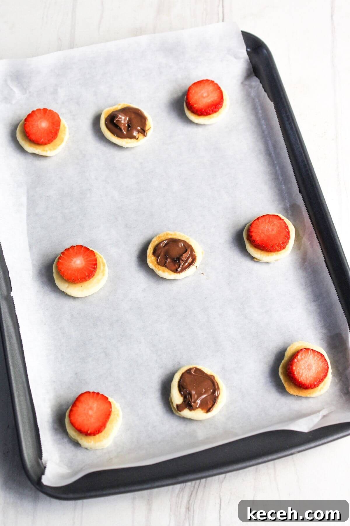 A baking sheet with eight mini pancakes, each topped with either a Nutella dollop or a strawberry slice, placed on parchment paper.