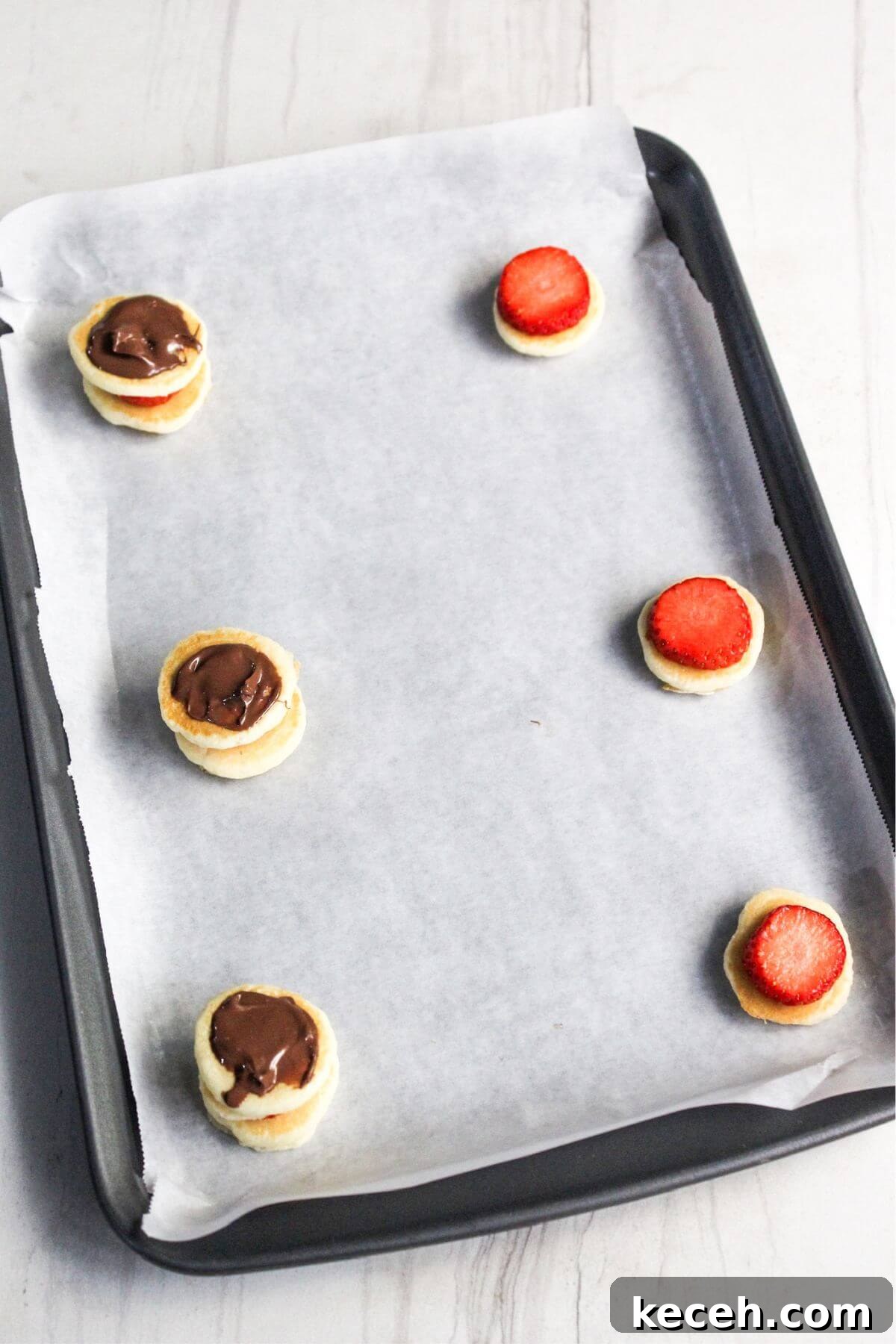 Baking tray with parchment paper, holding four mini pancake sandwiches: two with chocolate spread and two topped with strawberry slices.