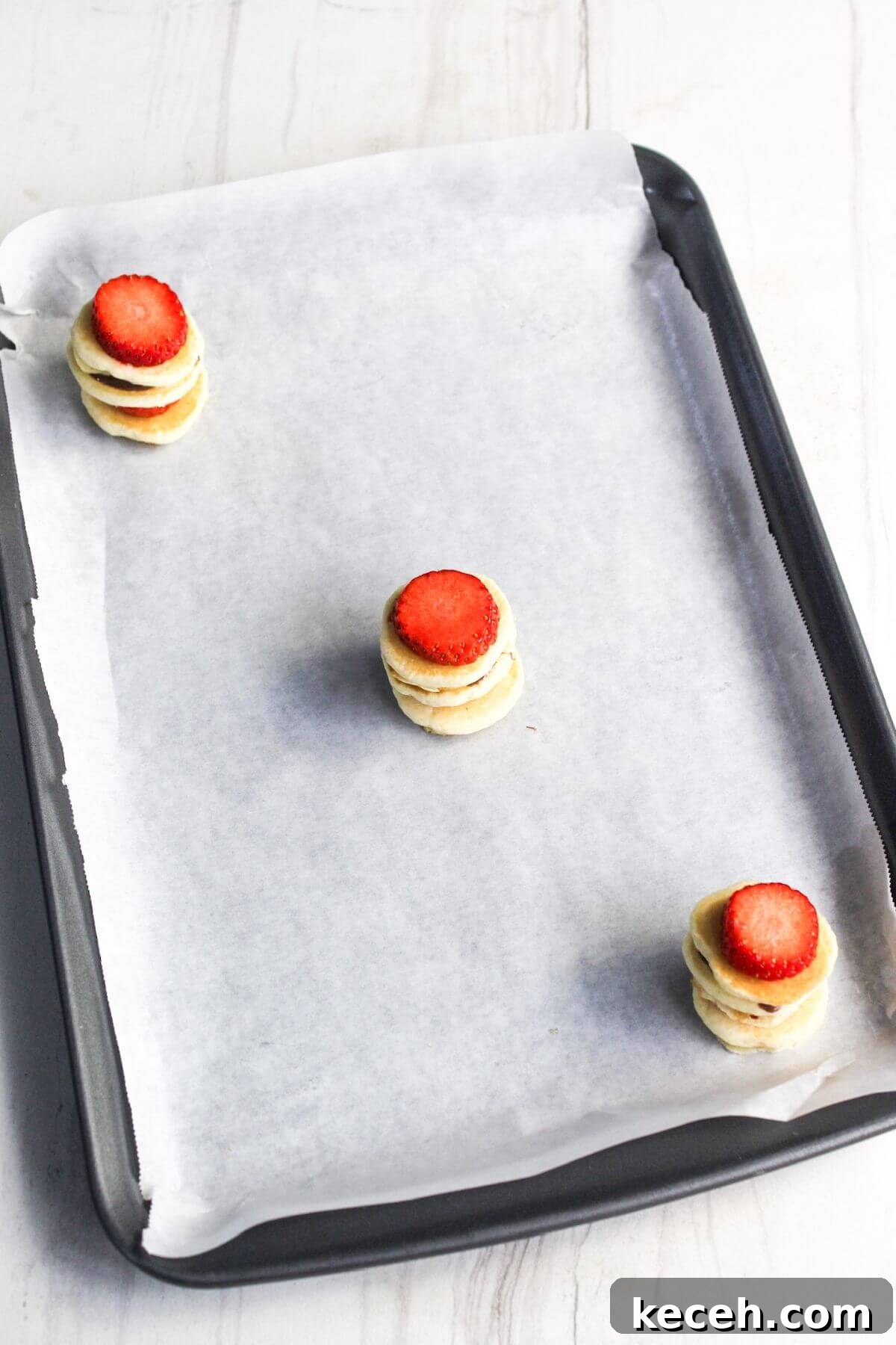 Three mini pancake stacks topped with strawberry slices are placed on a parchment-lined baking sheet.