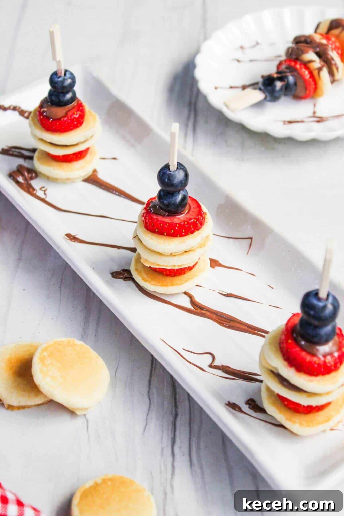 Three skewer sticks alternating with mini pancakes, strawberries, and blueberries on a white rectangular plate drizzled with chocolate. Nearby, a white dish holds another similar skewer and scattered mini pancakes.