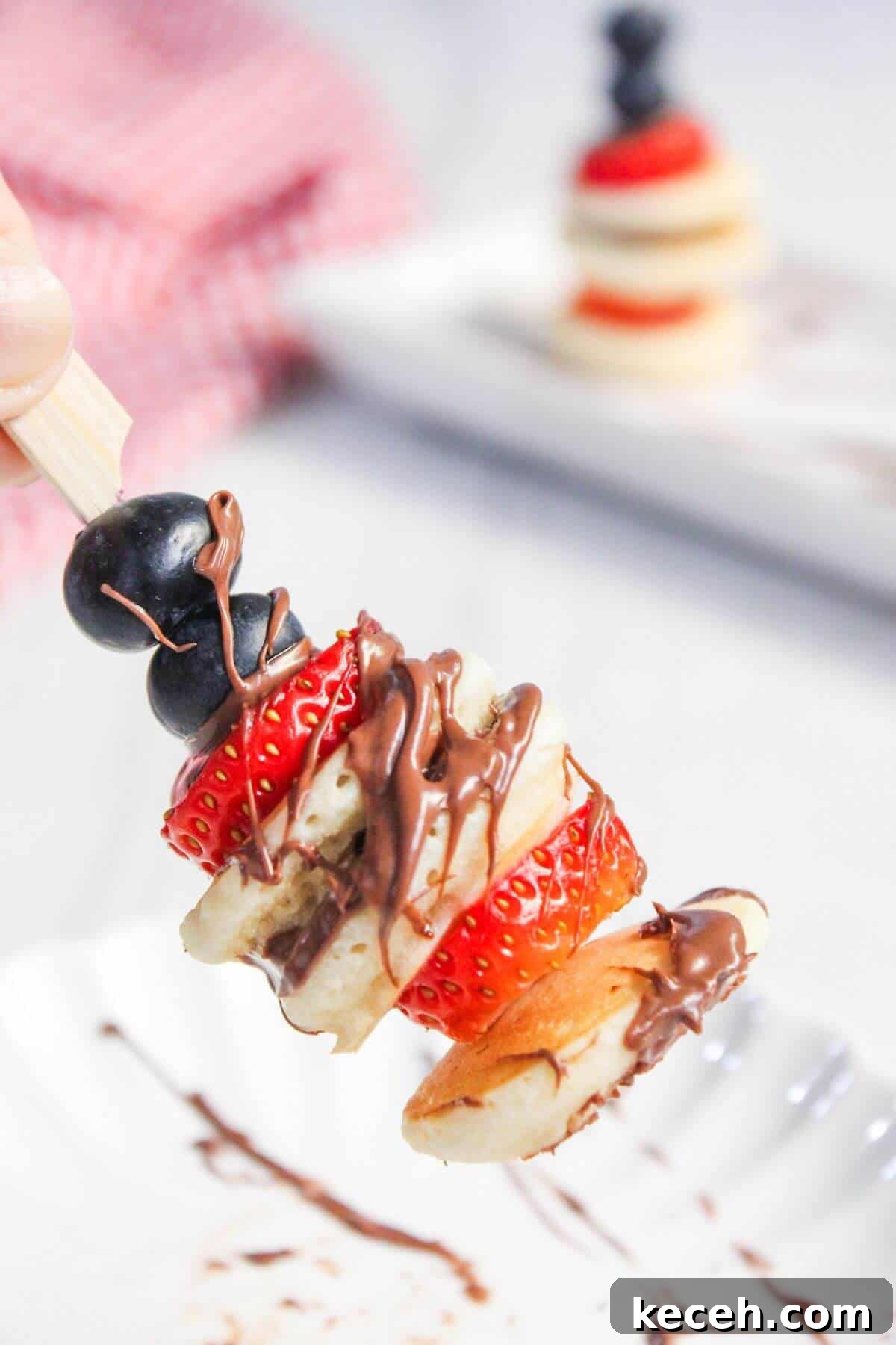 A skewer with stacked mini pancakes, strawberries, and blueberries drizzled with chocolate. A similar skewer is blurred in the background.