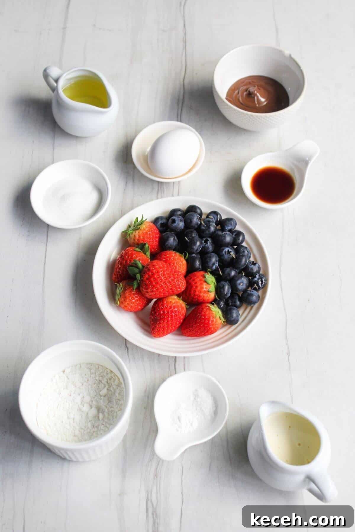 A set of ingredients including strawberries, blueberries, an egg, vanilla extract, chocolate spread, sugar, flour, oil, and cream arranged in bowls on a white surface.