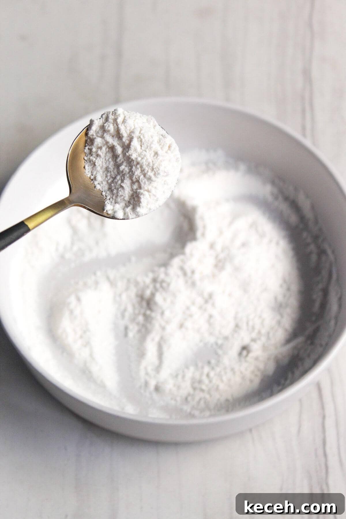 A spoon filled with flour mixture held over a white bowl containing the same white flour mixture, on a light-colored surface.