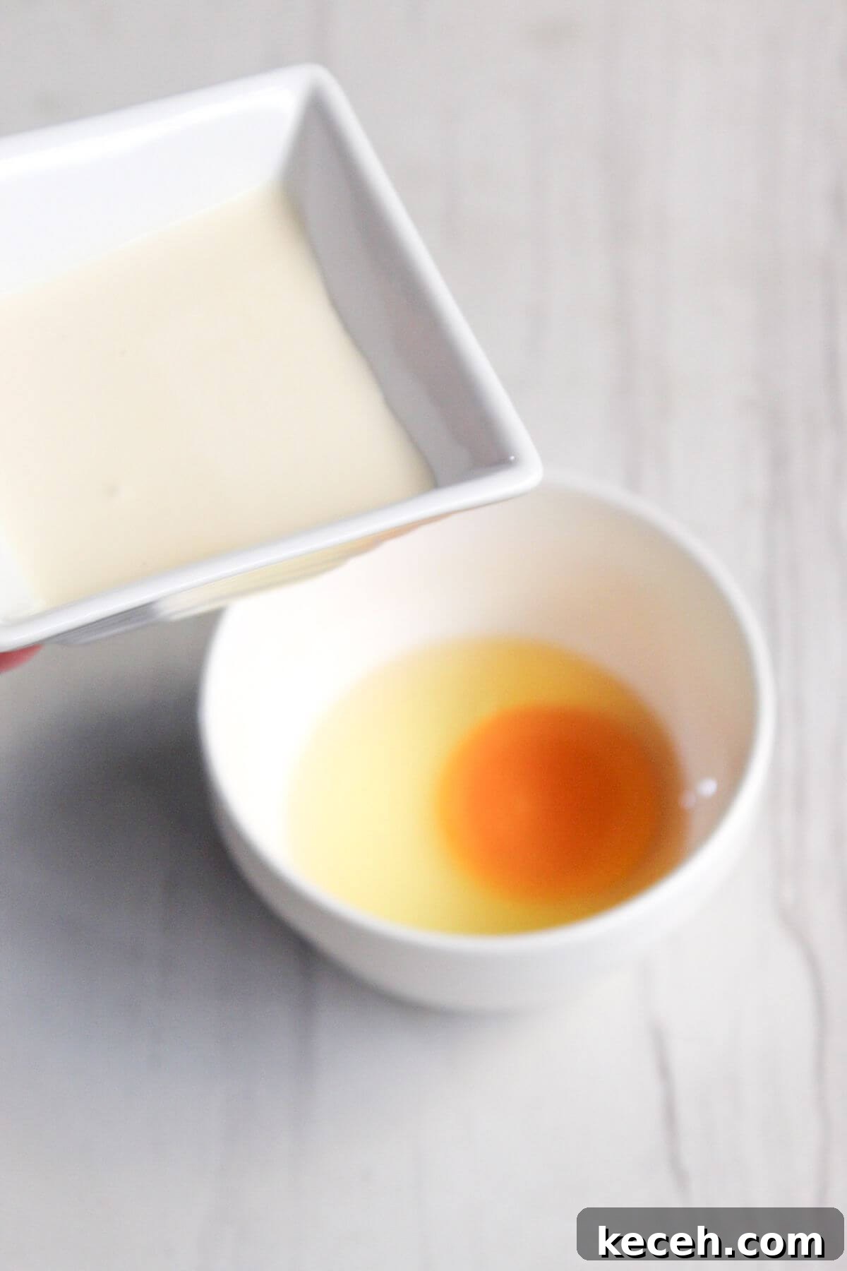 A square dish with cream is being held above a round bowl containing a raw egg yolk and white.