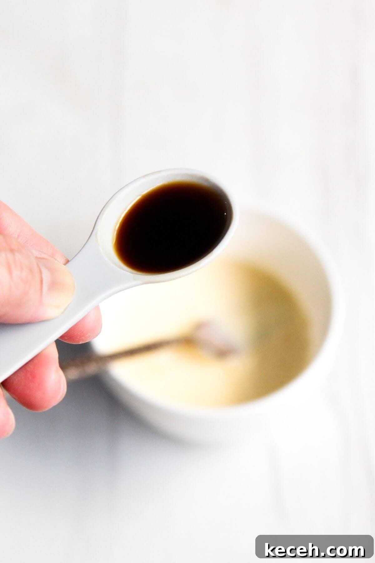 A hand holds a measuring spoon with vanilla extract over a white bowl containing a light mixture, with a spoon inside the bowl.