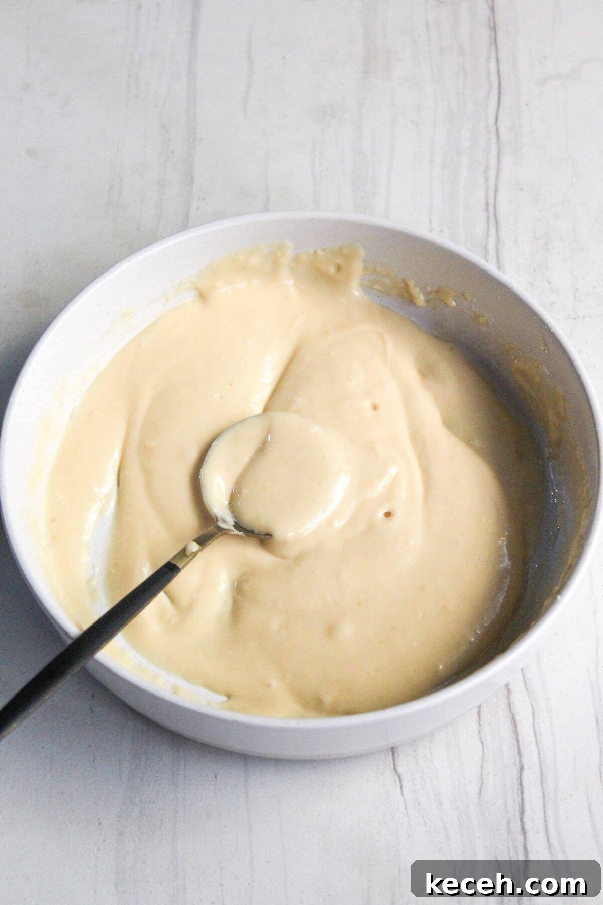 A bowl of thick, pancake batter with a spoon resting inside on a light surface.