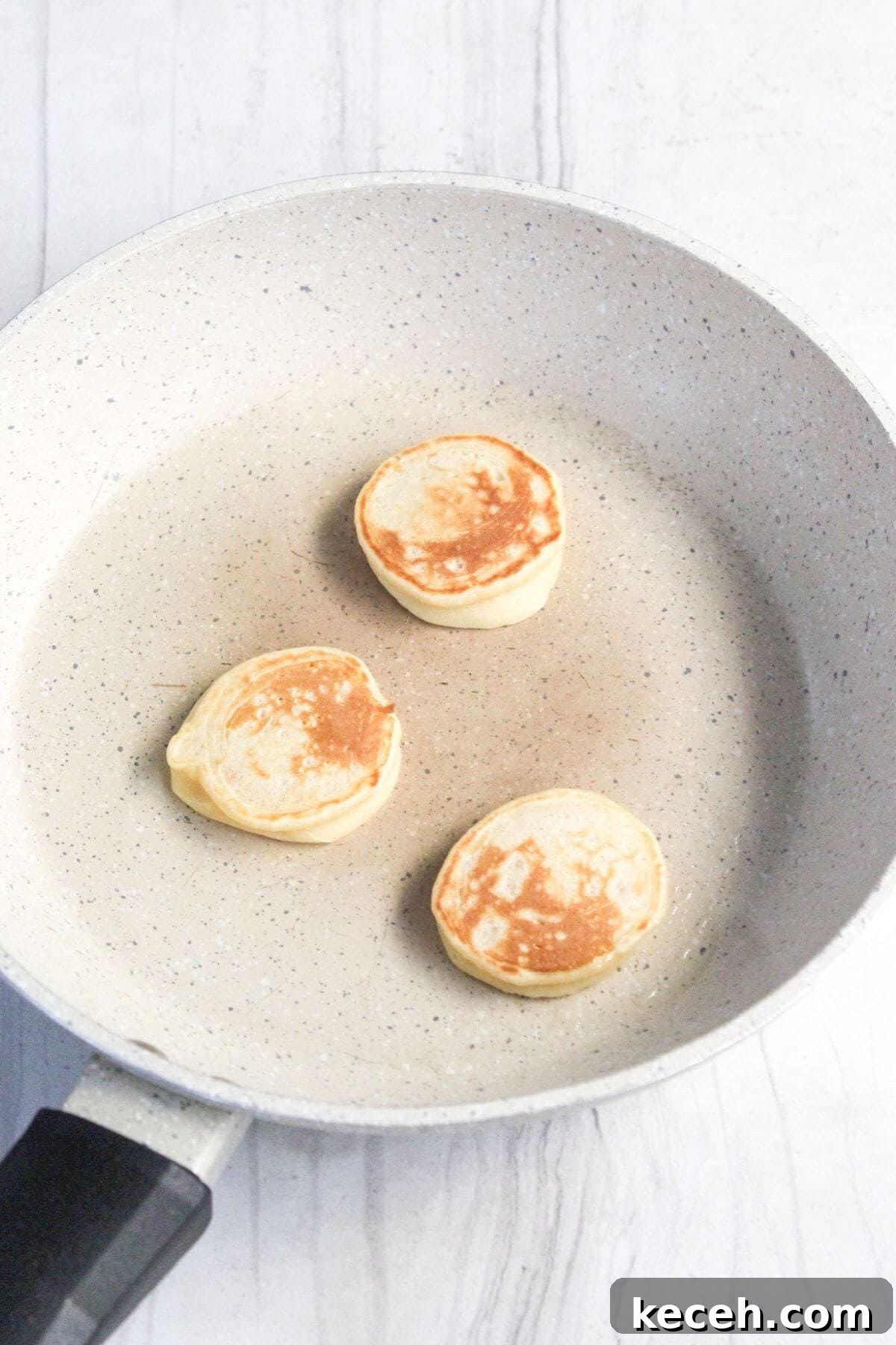 Three mini pancakes cooking in a non-stick frying pan on a white surface.