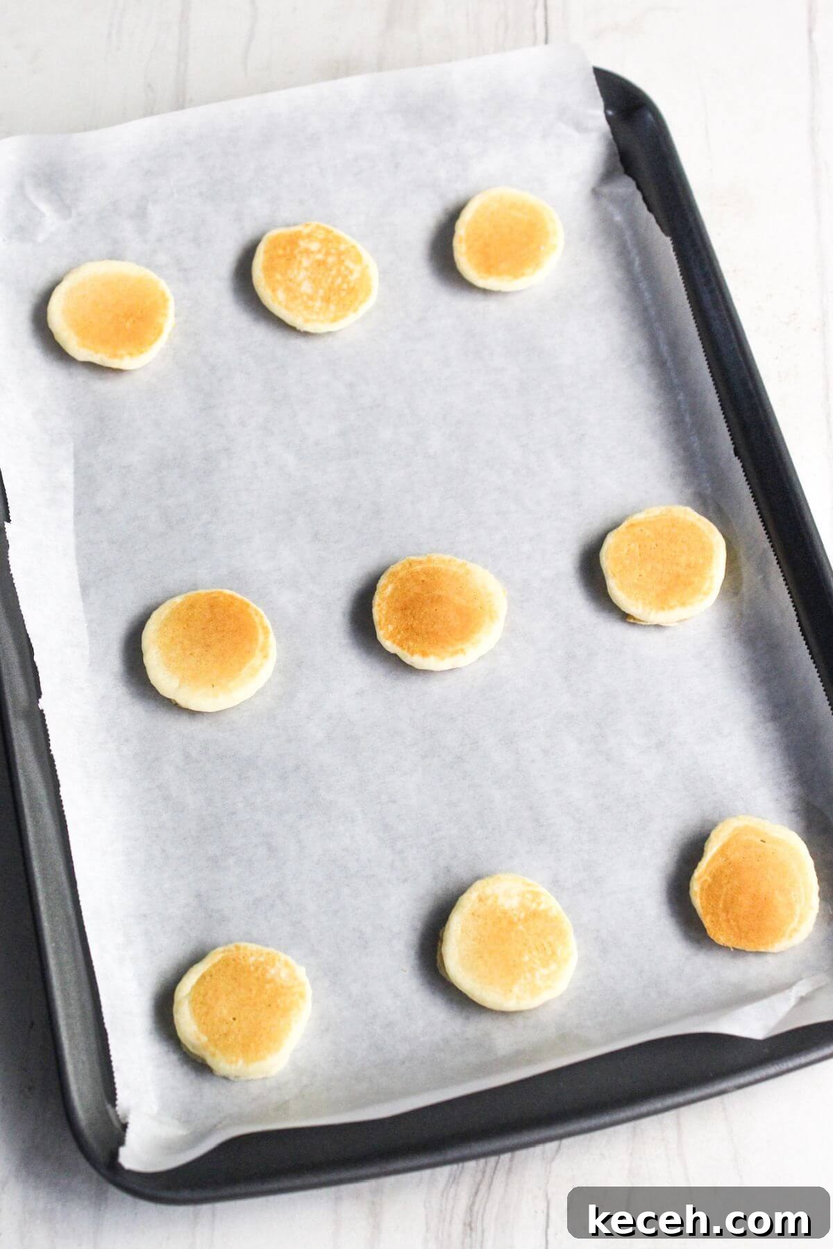 A baking tray lined with parchment paper holds nine mini pancakes spaced evenly.