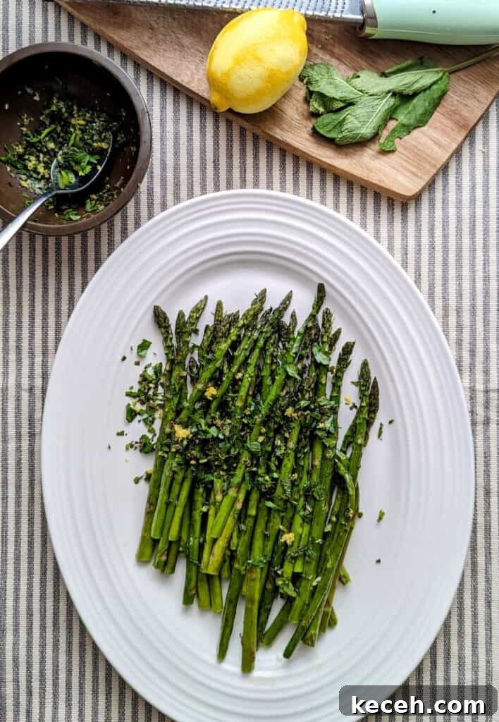 Roasted Asparagus with Fresh Mint Gremolata Recipe 2 Overhead tablescape shot of a platter of roasted asparagus topped with herbs and lemon zest on a white platter with a brown cutting board off center. The cutting board has a microplane with a lemon plus mint leaves. Off center is a small brown bowl with the mint gremolata.