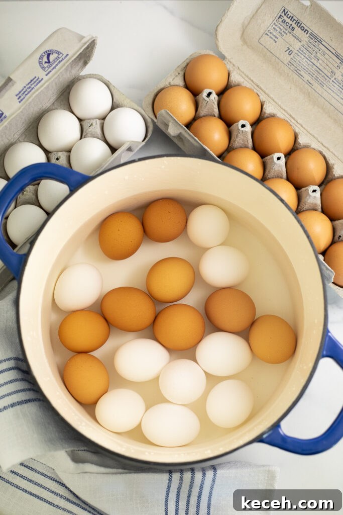 An overhead of a ceramic pot with 18 eggs, half white and half brown, covered with water with 2 cartons of eggs behind and a linen napkin next to the pot.