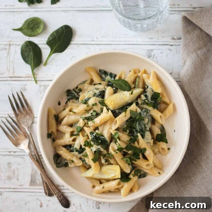 Bowl of vegan spinach artichoke pasta in a white bowl next to two forks.