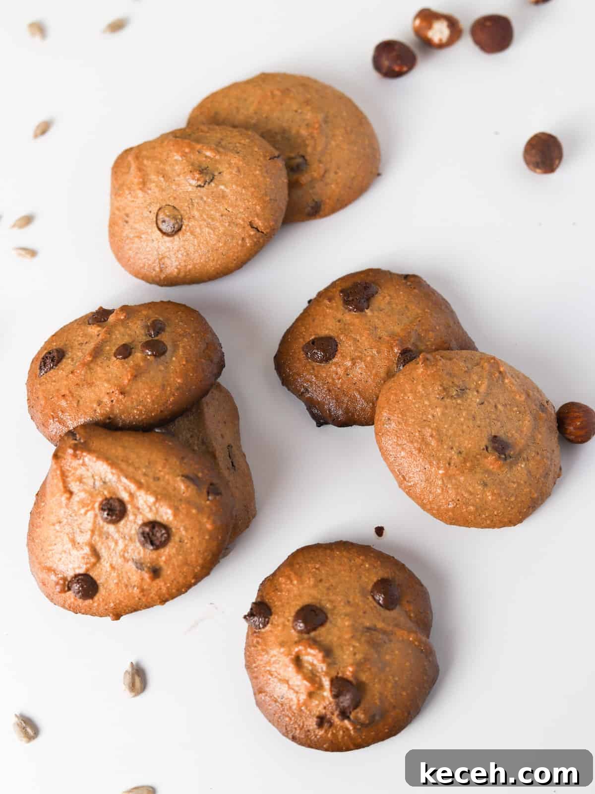 Photo of piles of cooked hazelnut and sunflower choc chip cookies on a white background with seeds and nuts scattered.