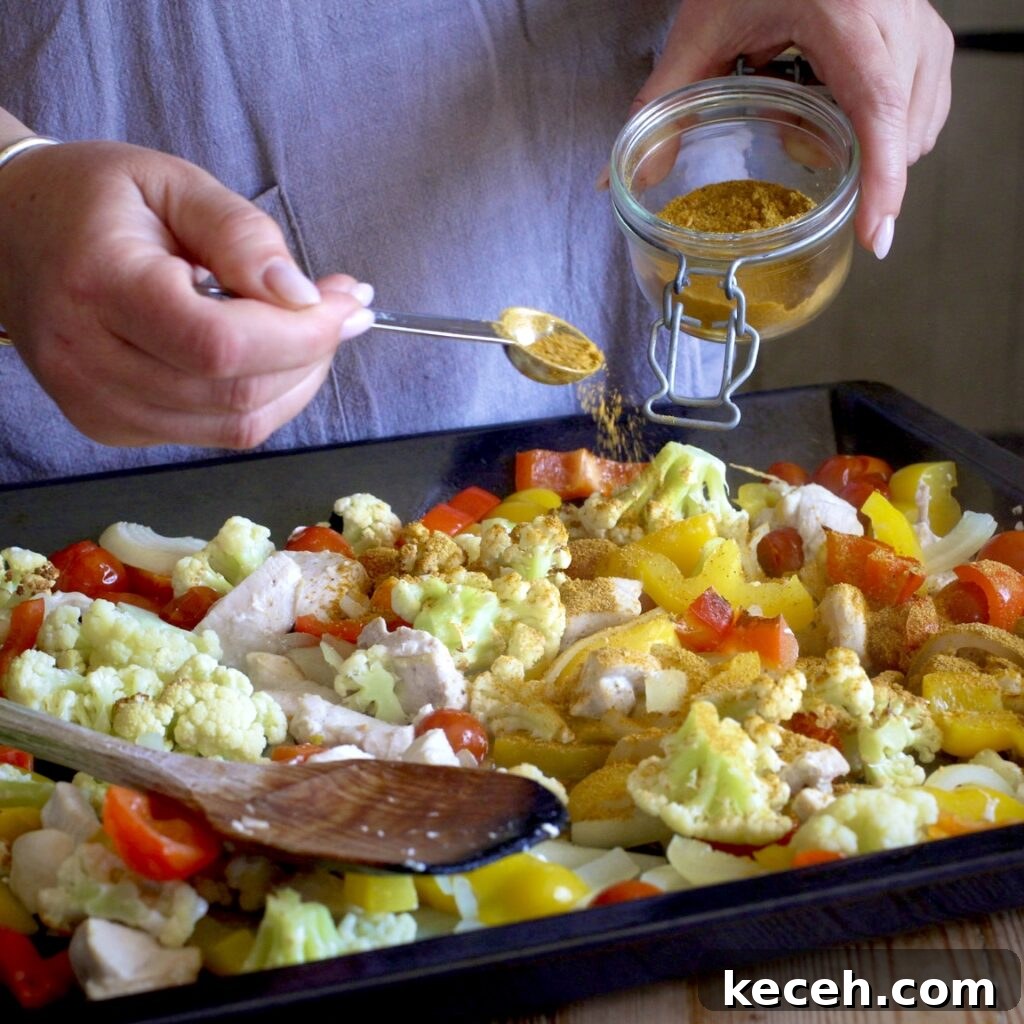 Curried Chicken Tray Bake Recipe from The Slimming Foodie 9 Woman in grey scattering a homemade spice blend over a baking tray of chopped veggies and chicken