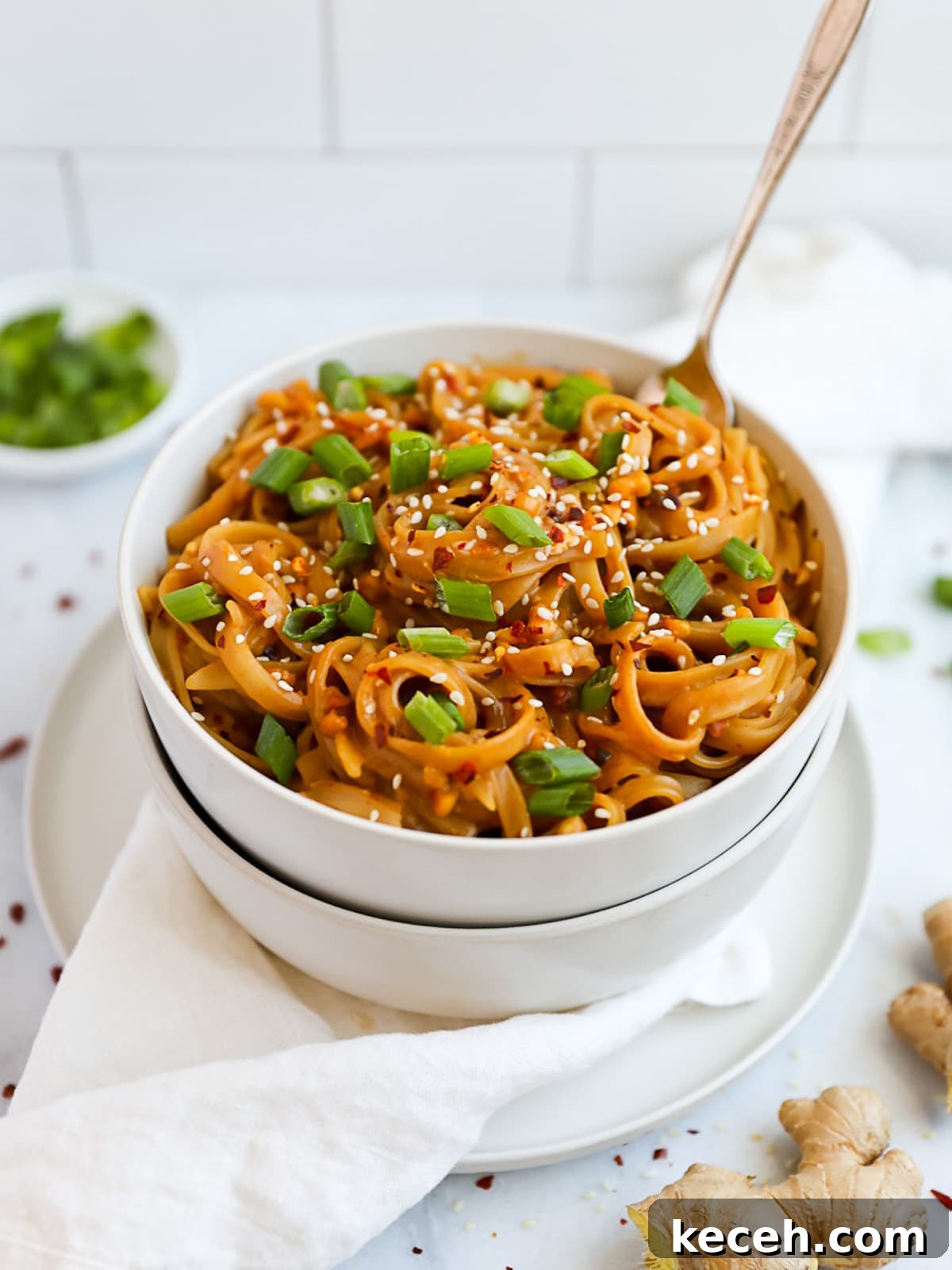Spicy Peanut Noodle Recipe: Creamy Chili Sesame Noodles 4 A white bowl with peanut noodles inside of it. It is garnished with green onions, sesame seeds, and crushed red pepper flakes.