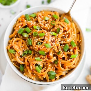 Spicy Peanut Noodle Recipe: Creamy Chili Sesame Noodles 6 A white bowl filled with creamy peanut butter noodles. They are garnished with green onions and white sesame seeds.