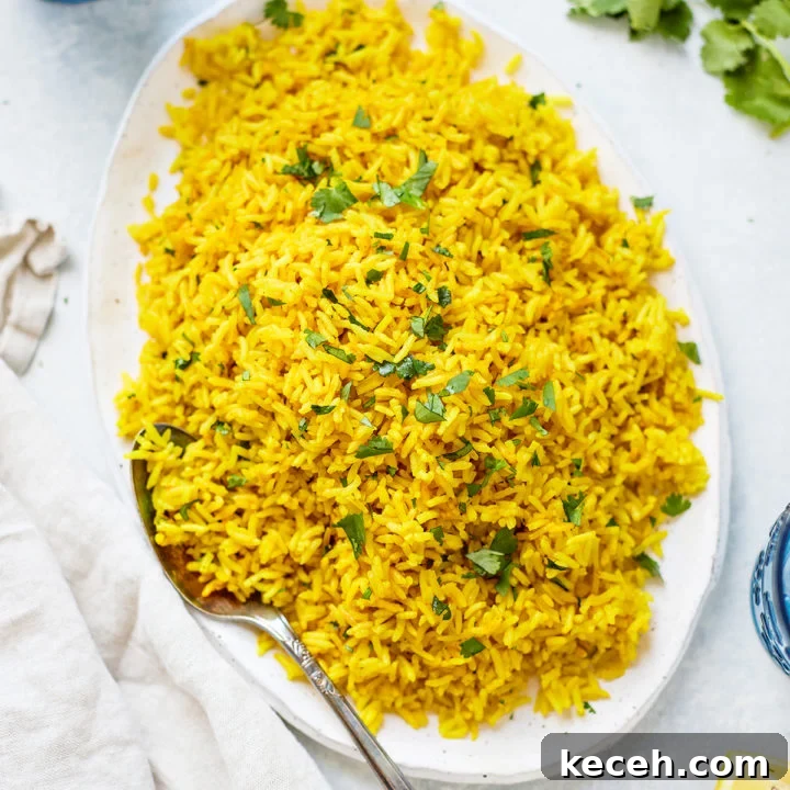 Turmeric yellow rice on an oval white platter, with fresh cilantro and a kitchen towel nearby. S spoon rests on the plate, ready to serve.
