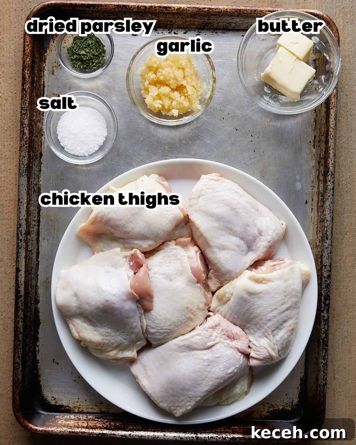 Crispy Baked Garlic Butter Chicken Thighs 3 Overhead shot of ingredients for garlic butter chicken thighs, including raw chicken thighs, butter, fresh garlic, and seasonings arranged on a baking sheet.