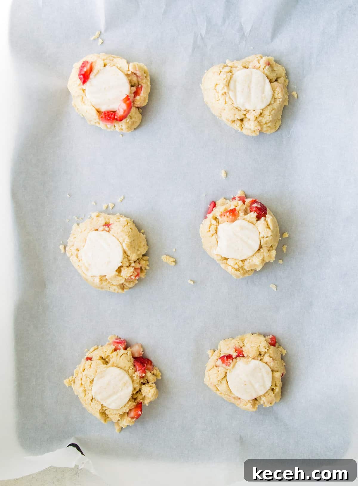 A piece of parchment paper with strawberry cookie dough balls on it with cheesecake pieces in each cookie.