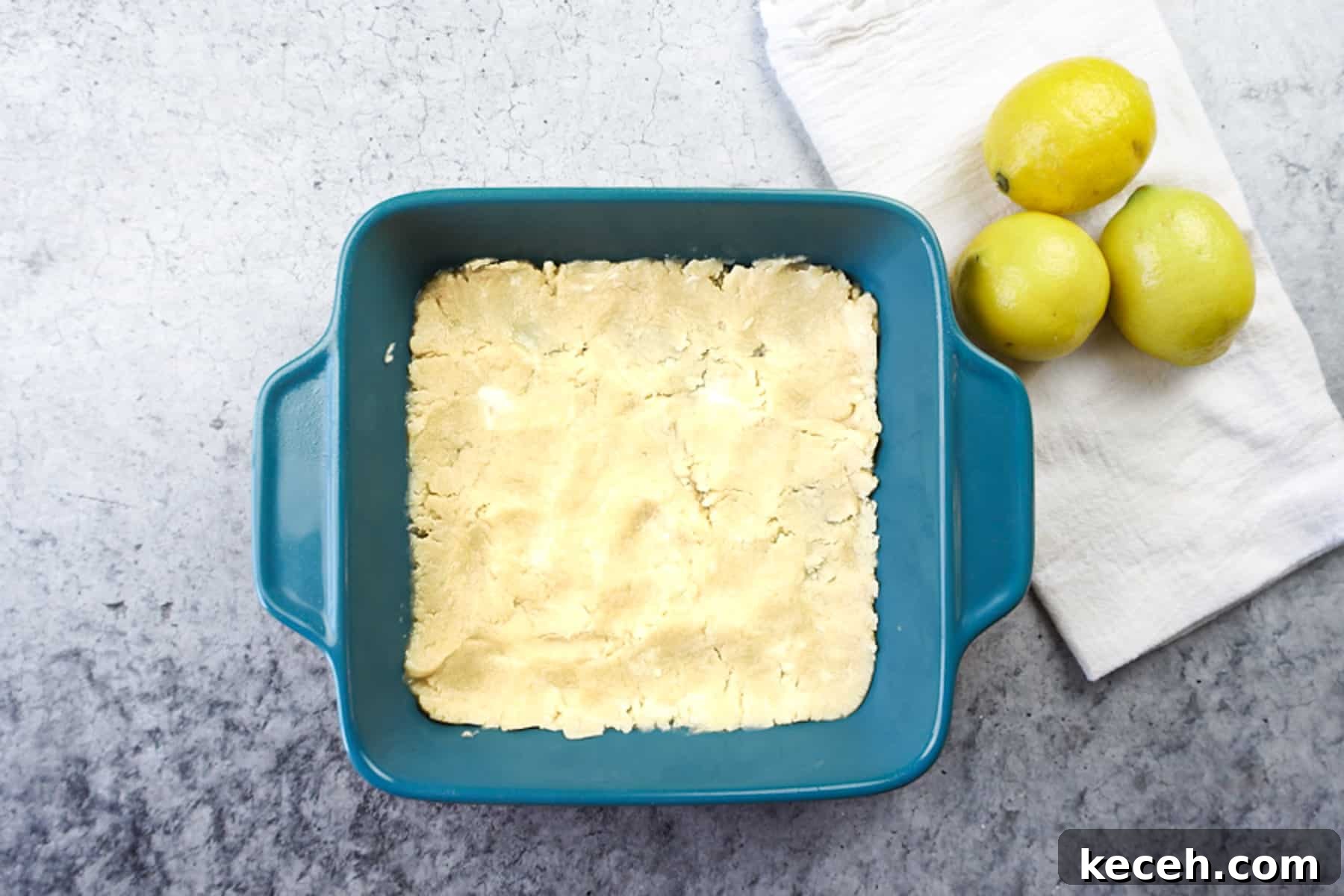 Classic Old-Fashioned Lemon Squares Recipe for Bright Dessert 6 shortbread crust pressed into teal baking dish next to a white towel with 3 lemons.
