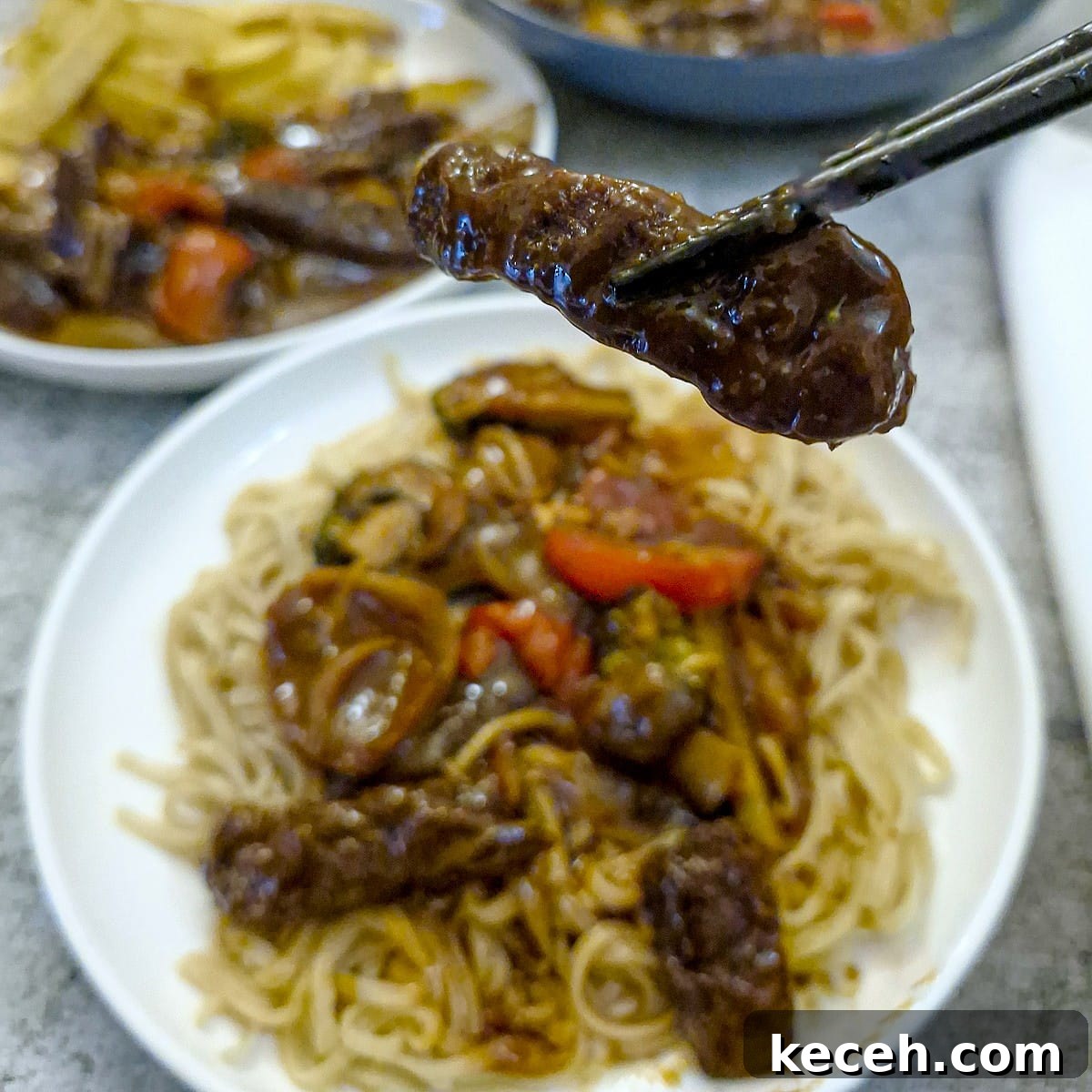 A piece of rump steak coated in garlic sauce, held in chopsticks above a plate of runp steak strips and noodles.