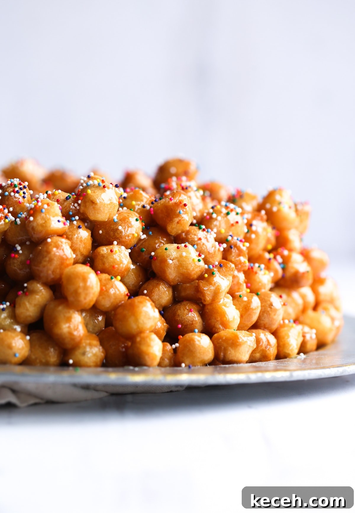 Authentic homemade Struffoli is such a fun Christmas dessert! Cute little balls of fried dough are coated in sweet honey and topped with your favorite holiday sprinkles. Struffoli piled on a serving platter topped with honey and sprinkles