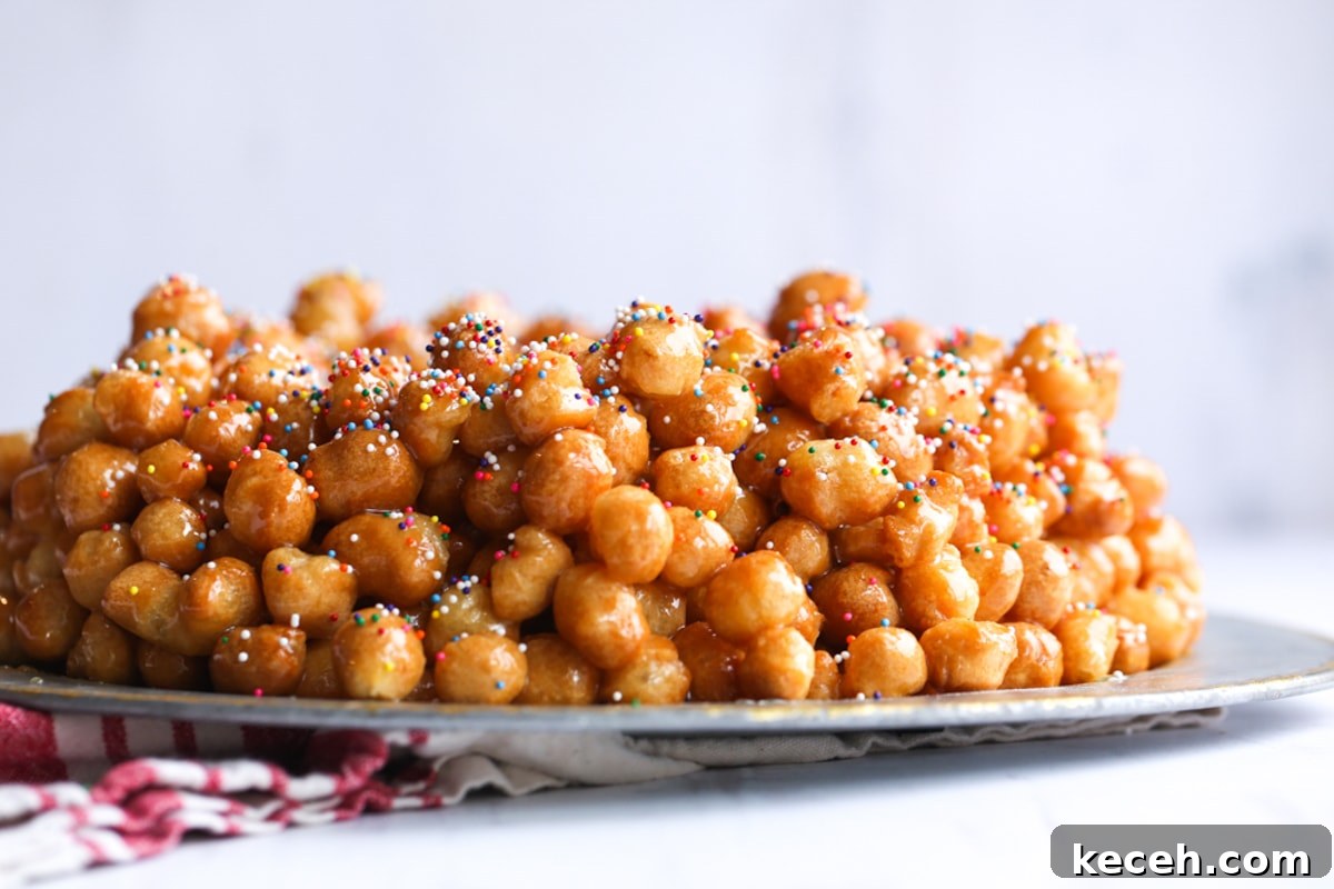 Authentic homemade Struffoli is such a fun Christmas dessert! Cute little balls of fried dough are coated in sweet honey and topped with your favorite holiday sprinkles. Struffoli honey balls on a silver serving plate topped with nonpareil sprinkles