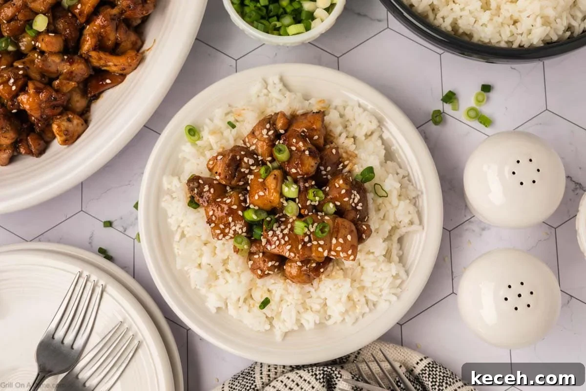 A plate of white rice topped with diced Blackstone Chicken teriyaki, sesame seeds, and chopped green onions. Surrounding the plate are a bowl of rice, a dish of chicken, salt and pepper shakers, forks, and a bowl of sliced green onions.
