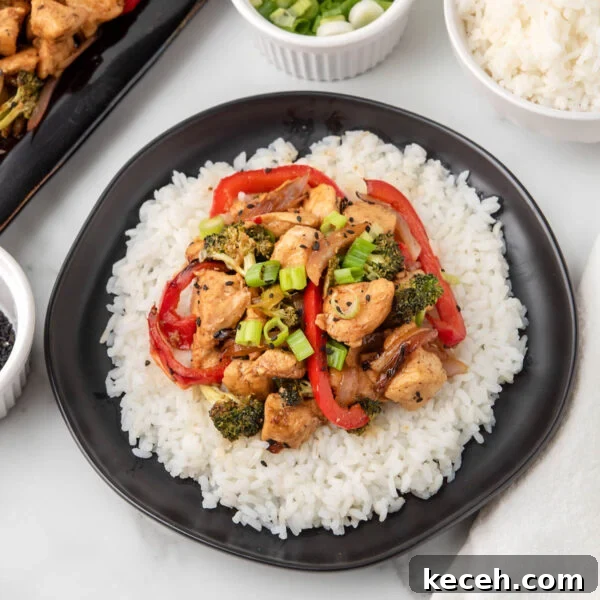 A black plate with white rice topped with Blackstone Teriyaki Chicken Stir Fry, featuring chicken, red bell peppers, broccoli, and green onions.