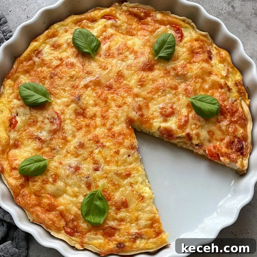 A delicious Chicken Quiche with a golden, cheesy top and a slice removed, garnished with five fresh basil leaves, sits in a round white ceramic dish on a gray surface.