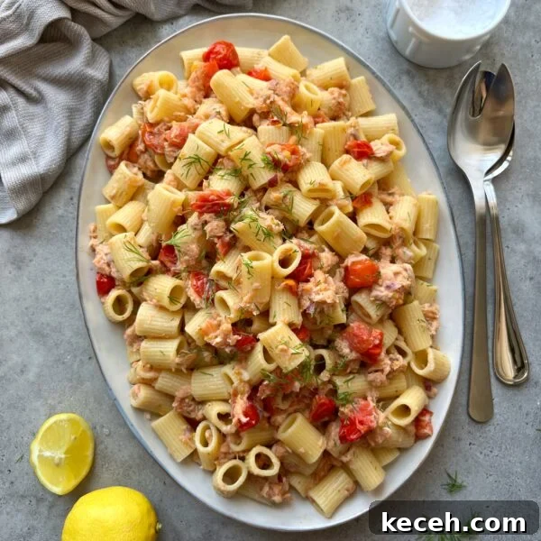 Oval platter of rigatoni pasta mixed with cherry tomatoes, salmon pieces, and fresh herbs, placed on a gray surface with a lemon, napkin, and utensils nearby.