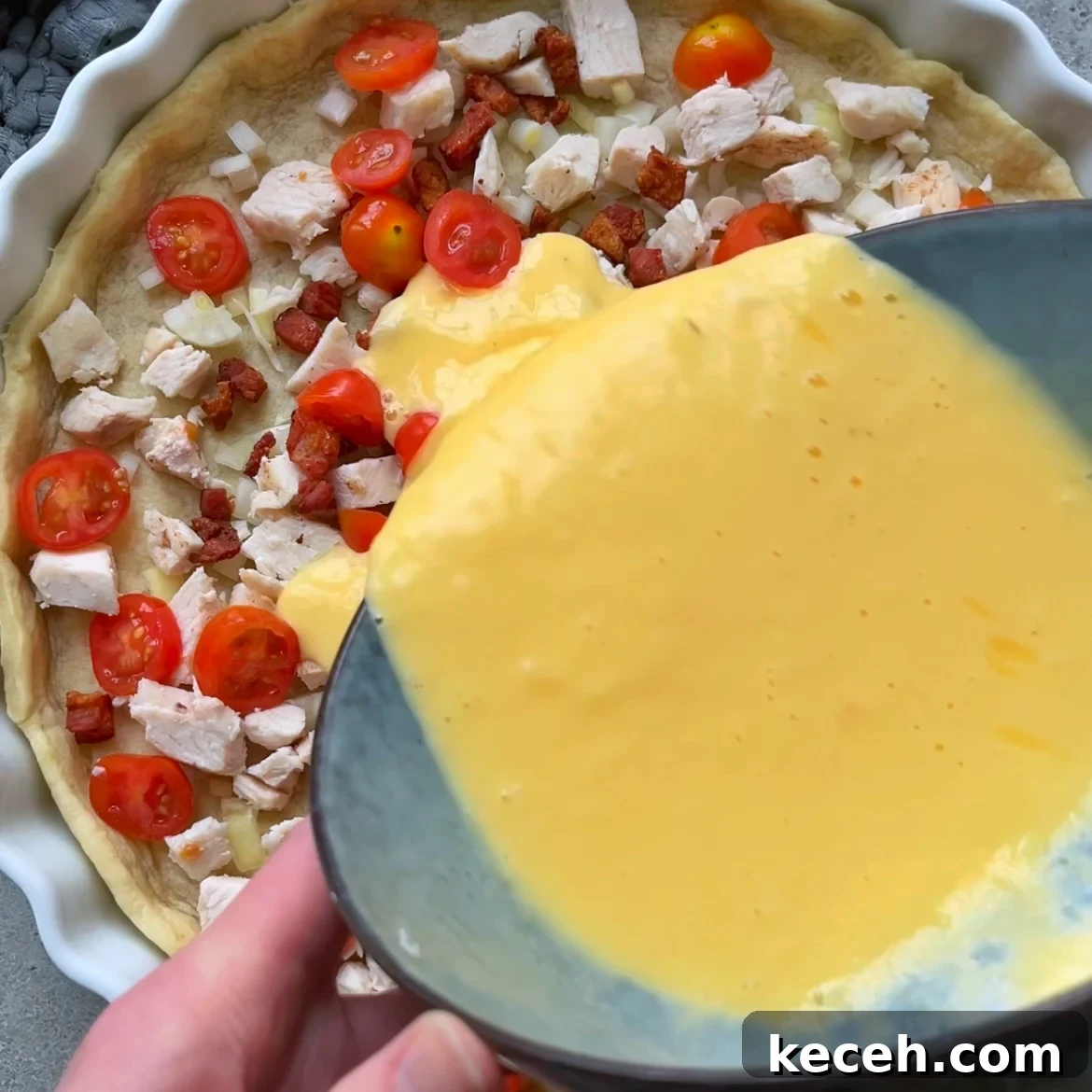 A bowl of beaten eggs is being poured over a pie crust filled with chopped chicken, cherry tomatoes, onions, and pancetta.
