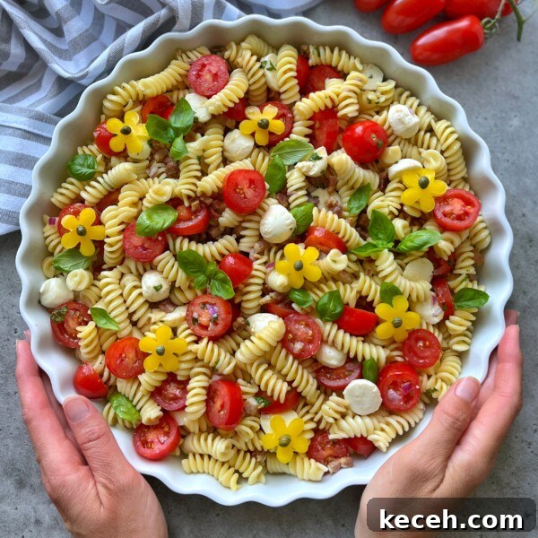 A person holds a round dish filled with healthy fusilli pasta salad, cherry tomatoes, mozzarella balls, basil leaves, and yellow flower-shaped bites, on a gray surface with grape tomatoes and a striped towel nearby.