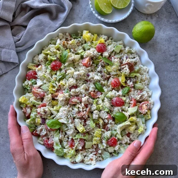 A person holds a round dish filled with a colorful rice chicken salad made of lettuce, cherry tomatoes, avocado, and fresh basil. Lime halves and a kitchen towel are nearby on a gray surface.