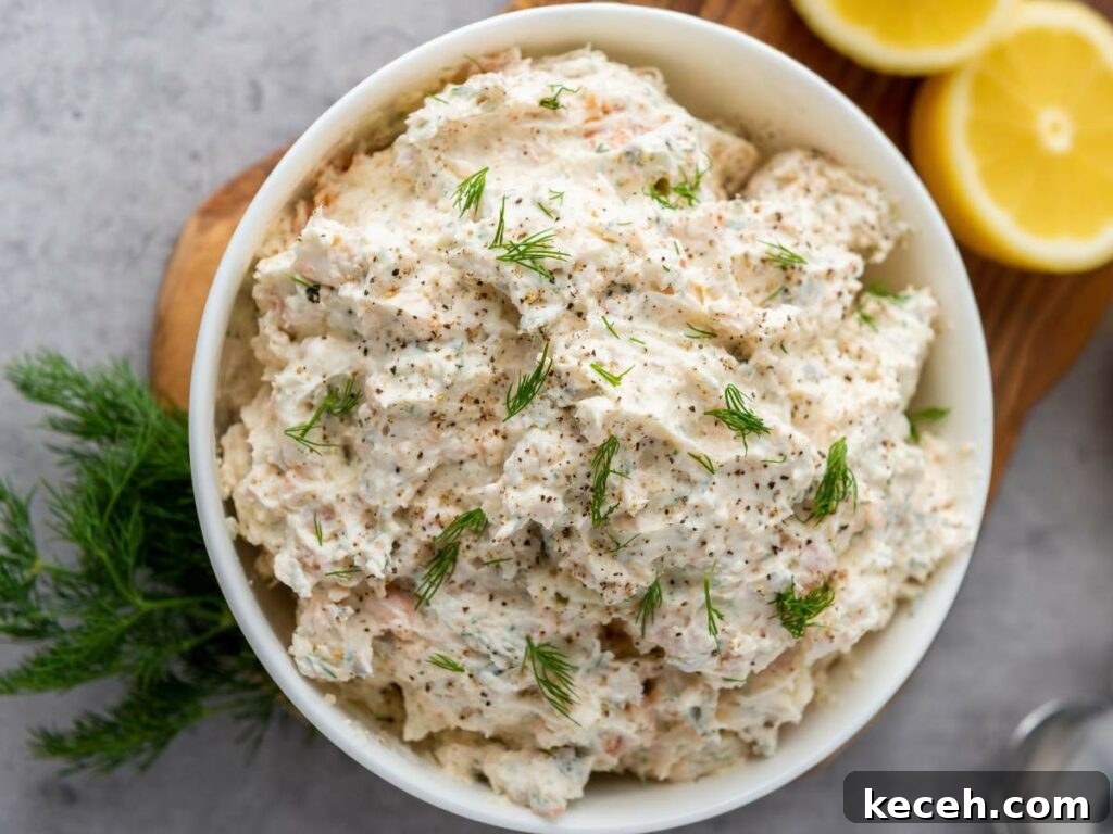 Bowl of salmon dip with cream cheese garnished with dill and pepper, placed on a wooden board.