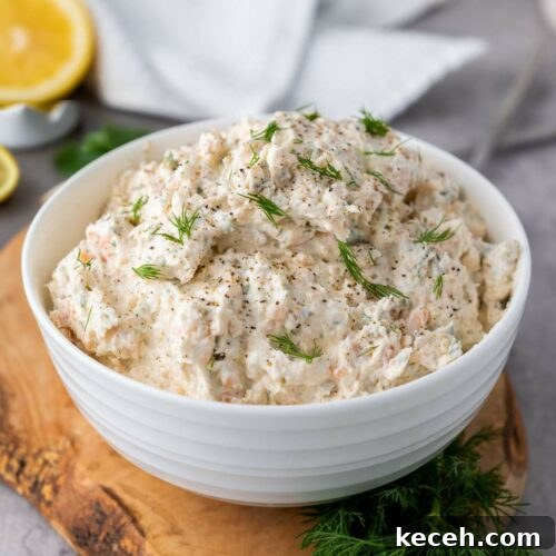 A white bowl filled with salmon dip with cream cheese garnished with dill on a wooden board.