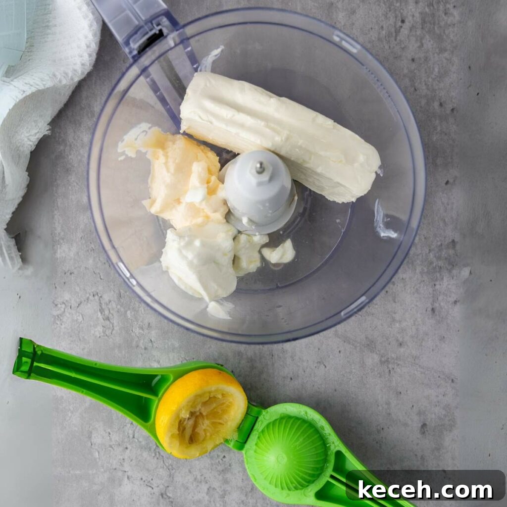 Food processor with cream cheese and mayo inside, next to a green citrus squeezer with a half-juiced lemon.