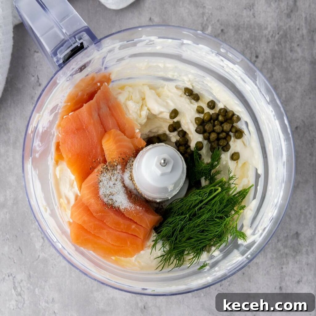 Food processor bowl with cream cheese, smoked salmon, capers, fresh dill, and seasoning, ready to be blended.
